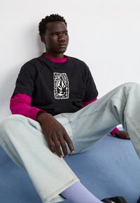 Camisa de algodón negra con un diseño gráfico blanco, superpuesta sobre una camiseta de manga larga rosa vibrante. Jeans de mezclilla azul claros, de corte holgado, combinados con zapatos oscuros.