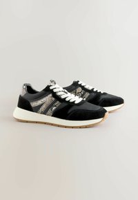 Melnas sporta kurpes ar zamša un čūskas rakstu akcentiem, baltām mežģīnēm un vieglu gumijas zoli. Iezīmē kontrastējošus materiālus un tekstūras.