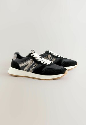 Melnas sporta kurpes ar zamša un čūskas rakstu akcentiem, baltām mežģīnēm un vieglu gumijas zoli. Iezīmē kontrastējošus materiālus un tekstūras.