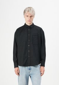 Chemise noire avec un motif à carreaux blancs, col boutonné, manches longues et poche poitrine. Le tissu semble lisse avec une coupe structurée.