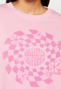 Rosa sweatshirt med en cirkulär grafisk design med rutmönster och blommor. Texten säger "HOLLISTER CALIFORNIA" i fet stil.
