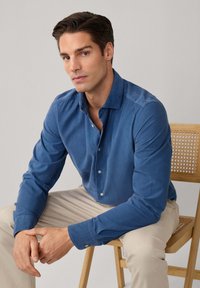 Camicetta in cotone blu solido, con colletto classico e maniche lunghe, abbinata a pantaloni beige. Tessuto morbido e design aderente.