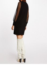 Robe noire avec manches longues en maille translucide texturée et coupe ample. Portée avec des bottes blanches montantes jusqu'aux genoux, dotées d'un talon noir épais.