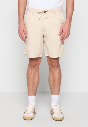 Moški, oblečen v bež kratke hlače z vrvico, bele športne nogavice in bež-bela superga, stoji na enobarvnem ozadju.