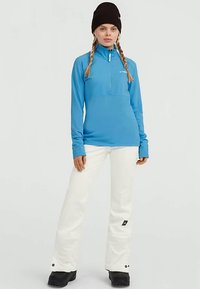 Woman with braided hair wearing a black beanie, blue quarter-zip jacket, white snow pants, and black boots standing against a plain background.