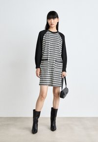 Mustavalkoinen houndstooth-mekko, jossa on pitkät mustat hihat, kaksi etutaskua mustilla yksityiskohdilla, yhdistettynä polven yli ulottuviin mustiin saappaisiin.