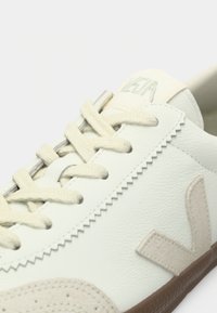 Bílá kožená teniska s béžovými semišovými doplňky a texturovanou hnědou gumovou podrážkou. Má ploché tkaničky a kontrastní švy.