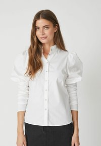 Camicia bianca con maniche a sbuffo e colletto arricciato. Realizzata in morbido tessuto di cotone. Presenta dettagli di bottoni discreti e polsini aderenti.