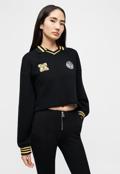 Μαύρο κοντό φούτερ με κίτρινο ριγέ γιακά, διακοσμημένο με δύο κεντημένα μπαλώματα. Μανσέτες με ριμπ ύφανση και τελείωμα με ακατέργαστο τελείωμα. Συνδυάζεται με μαύρο παντελόνι.