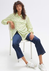 Light green button-up shirt with a wave pattern, paired with dark blue flared jeans and white sneakers, seated on a white chair.