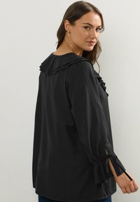 Svart blus med röflat krage och ärmar med band. Tillverkad av lätt tyg med en slät textur. Enkel, klassisk design.