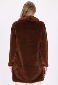 Cappotto in finta pelliccia marrone e morbido, con colletto, maniche lunghe e taglio dritto che arriva sotto i fianchi. Texture liscia e pelosa.