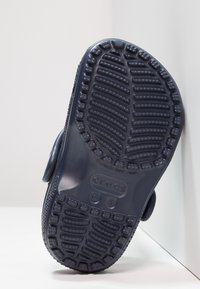 Crocs blu navy con suola in gomma testurizzata. Presentano un grande bottone circolare e scanalature per la trazione. Design piatto, materiale stampato.