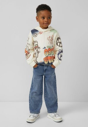 Cremefarbiger Hoodie mit bunten Graffiti-Drucken, einschließlich orangefarbener Schriftzüge, kombiniert mit lockeren blauen Jeans und weißen Sneakers.