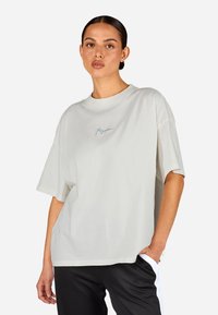 Világosszürke, oversize pamut póló, kerek nyakkal és rövid ujjal, a mellkasán egy diszkrét kék logóval.