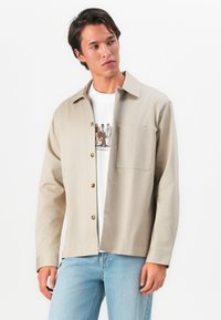 Young man with dark hair wearing a beige button-up jacket over a white graphic t-shirt and light blue jeans, looking to the side against white background.