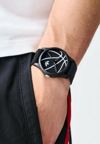 Montre noire avec un boîtier rond, bracelet texturé noir et graphique de basket-ball blanc sur le cadran ; couronne argentée sur le côté.