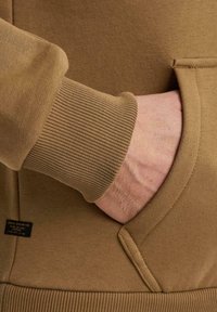 Brown sweatshirt material with ribbed cuffs and hem. Features a kangaroo pocket and a small black tag on the lower left side.