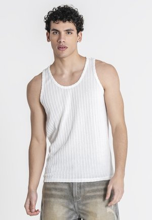 Junger Mann mit lockigem dunklem Haar, der ein weißes geripptes Tanktop und ausgewaschene graue Jeans trägt, steht vor einem schlichten hellen Hintergrund.