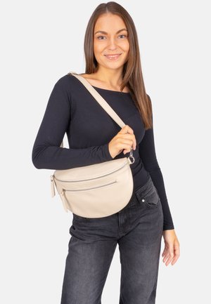 Femme aux longs cheveux bruns portant un haut noir à manches longues et un jean foncé, tenant un sac bandoulière beige sur sa poitrine.