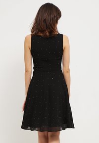 Musta hihaton mekko, jossa tiimalasityylinen yläosa ja liehuva helma, koristeltu hohtavilla hopeisilla pisteillä kauttaaltaan. Sileä, kevyt kangas.