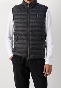 Hombre que lleva un chaleco sin mangas negro acolchado con cremallera frontal y bolsillos con cremallera sobre una camisa de manga larga blanca y pantalones negros.