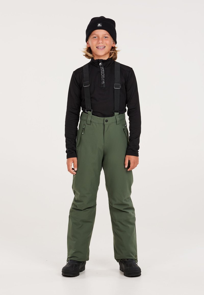 Groene sneeuwbroeken met verstelbare zwarte bretels, zijzakken met rits en een versterkte taille, gecombineerd met een zwart languit T-shirt en een zwarte muts.