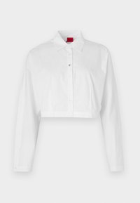 Camicia bianca cropped con maniche lunghe, dotata di colletto classico e dettagli del logo discreti vicino ai bottoni. Texture liscia.
