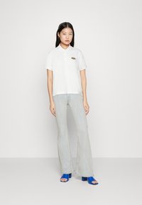 Camisa de polo blanca con mangas cortas y cuello, que presenta un logo en verde y rojo. Jeans acampanados de un azul claro y sandalias de tacón bajo en azul.