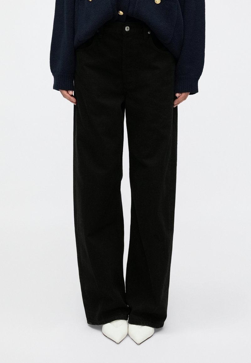 Čierne široké nohavice z hrubej látky s vysokým pásom, prednými vreckami a jemným zapínaním na gombík. Topánky sú biele a špicaté.