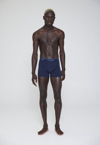 Boxer briefs in cotone blu navy con una vita blu a contrasto con l'etichetta "CLAUDIO," indossati da un modello, in piedi contro uno sfondo neutro.