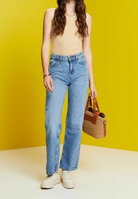 Jeans de corte recto de talle alto en azul claro, combinados con una camiseta sin mangas beige y zapatillas blancas. Sosteniendo un bolso de paja color marrón.
