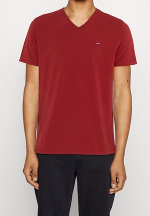 Mand iført en ensfarvet rød Levi's T-shirt med V-udskæring og korte ærmer samt sorte bukser, stående foran en ensfarvet baggrund.