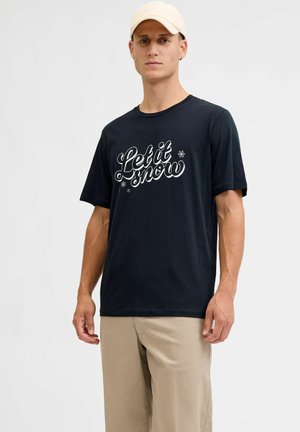 Zwarte katoenen t-shirt met witte scripttekst "Let it snow" en sneeuwvlokaccenten; gedragen met lichtbeige broeken.