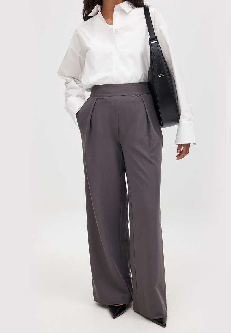 Grijze wijde broek gemaakt van een gladde stof, met een hoge taille, voorplooien en zijzakken, gecombineerd met een wit shirt.