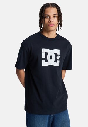 Jonge man met gevlochten haar, gekleed in een zwart T-shirt met een groot wit DC-logo en een blauwe spijkerbroek, staand tegen een effen achtergrond.