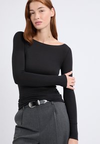 Schwarzes Langarmshirt mit einem Bootsausschnitt und figurbetontem Schnitt, kombiniert mit grauen, maßgeschneiderten Hosen und einem schwarzen Gürtel mit silberner Schnalle.