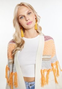 Mönstrad stickad cardigan i flera färger med sektioner i orange, grått och krämfärgade med fransdetaljer. Kombinerad med en vit topp och gula tofsörhängen.