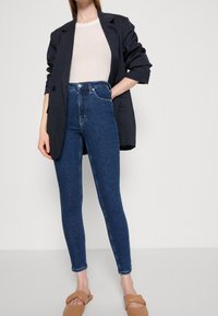 Tmavě modré džíny s vysokým pasem, slim fit, spárované s béžovým topem a černým sakem, s jemným elastickým materiálem a klasickým pětikomorovým designem.