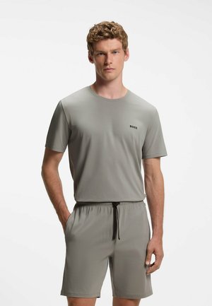 Mand med krøllet hår iført en grå kortærmet skjorte og matchende shorts med sort snøre, stående med den ene hånd i lommen.