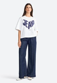 Chemise blanche à manches courtes avec le texte "JEANS" en bleu vif, associée à un pantalon en denim foncé à jambes larges et des chaussures blanches à bout pointu.