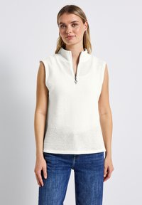Weißes, ärmelloses Oberteil mit hohem Kragen und Reißverschlussdetails. Hergestellt aus strukturiertem Stoff mit lockerer Passform. Kombiniert mit blauen Denim-Hosen.