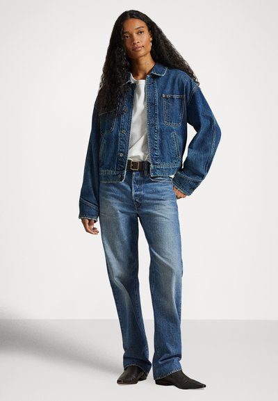 Kvinne med langt krøllet hår iført blå denimjakke, hvit skjorte, blå jeans, svart belte og svarte spisse sko som står foran en hvit bakgrunn.