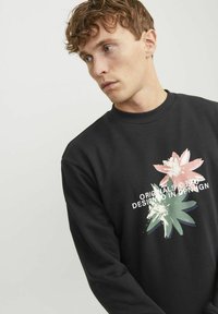 Sudadera negra con cuello redondo, que presenta un diseño floral en rosa y verde con texto blanco que dice "ORIGINALS © 1990 DISEÑADO EN CPH."