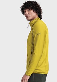 Gelbe Jacke mit Reißverschluss und einem strukturierten, gerippten Design. Verfügt über Seitentaschen und ein Logo am Oberarm. Getragen mit dunkler Hose.