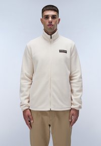 Creme fleecejakke med høj krave, fuld lynlås og et lille logo patch på brystet. Glat tekstur, tætsiddende silhuet og lange ærmer.