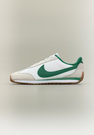 Baskets Nike blanches et beiges avec un swoosh et un col verts, semelle en gomme, affichées de profil sur un fond neutre.