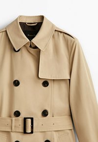 Trench-coat beige à double boutonnage avec boutons noirs, taille ceinturée, épaulettes et large col à revers.