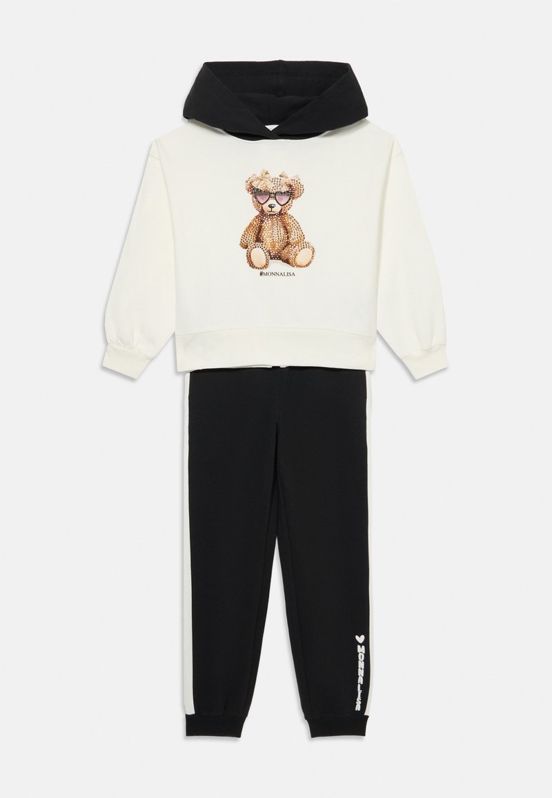 Sweat à capuche beige avec des accents noirs, featuring un motif de ours en strass ; joggeurs noirs avec du texte blanc sur le côté.