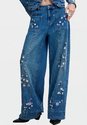 Vaqueros azules de pierna ancha con bolsillos delanteros de parche y bordado floral en púrpura y blanco, llevados con zapatos negros puntiagudos.